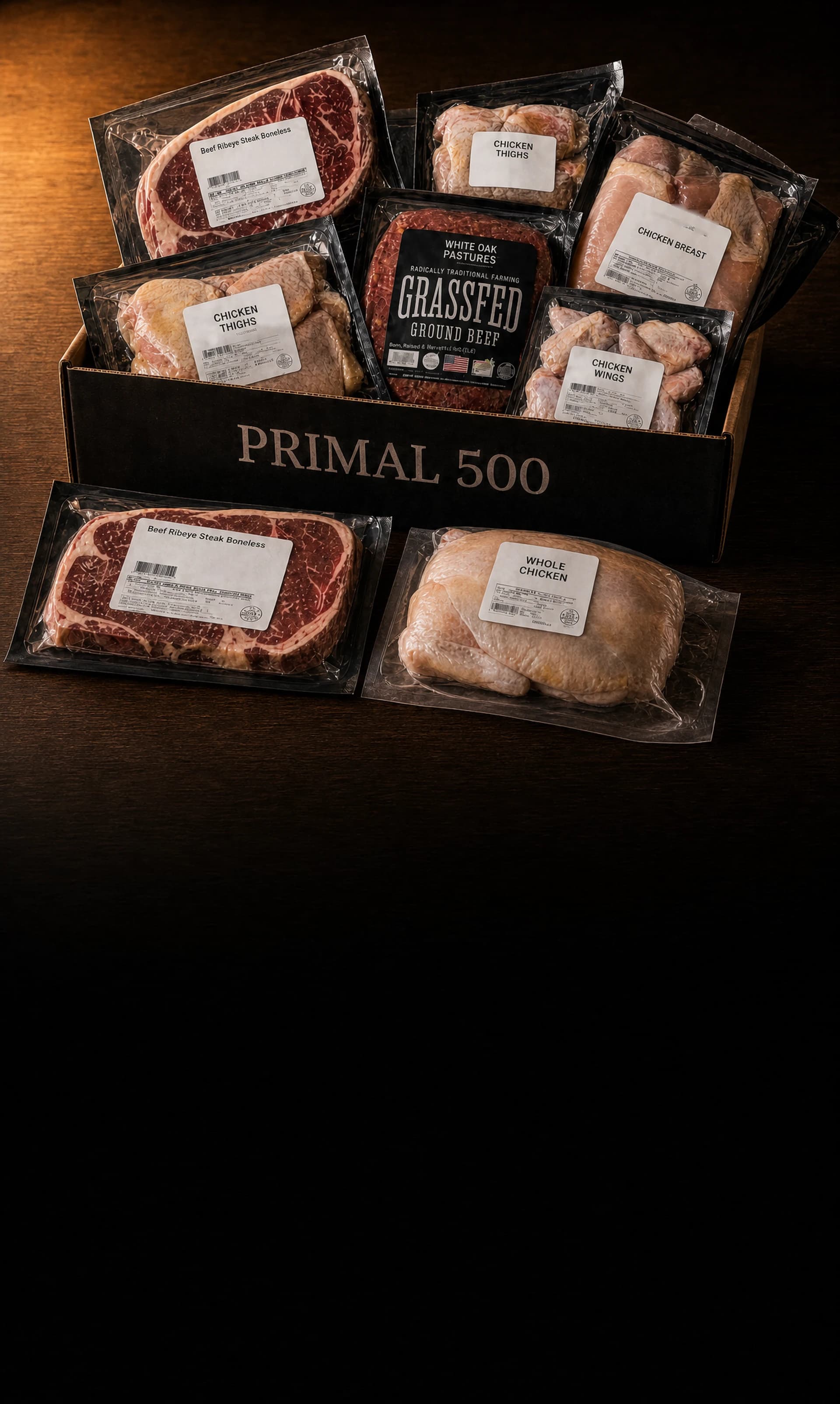 The Primal 500 Founding Member Box on a wooden farmhouse table, packed with labelled vacuum packs: ribeye steaks, White Oak Pastures grass-fed ground beef, chicken breast, thighs and wings, with a second ribeye and a whole chicken resting on the table in front of the box.